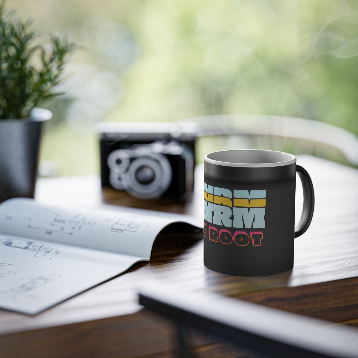 WNRM The Root Logo Magic Mug, 11oz, on a desk next to a camera and plant by a window