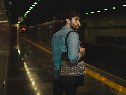 Good looking man carrying a WNRM Name In Lights canvass tote bag, while wearing a jean jacket, waiting for the subway 