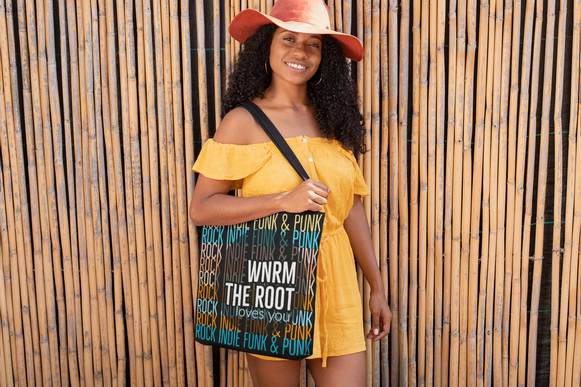 A pretty woman, dark hair, yellow romper. Holding a WNRM Loves You canvass bag by a bamboo fence.  